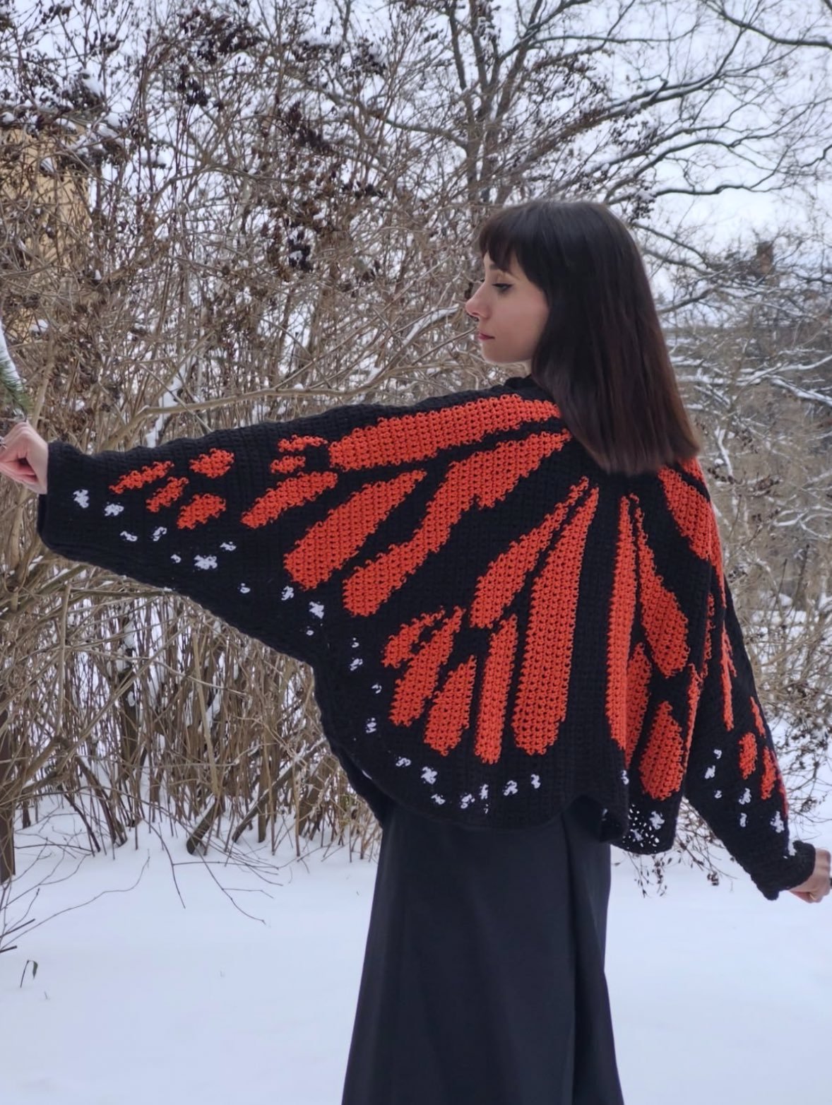 A hand-knitted cardigan patterned as monarch butterfly wings, worn with arms spread in the snow